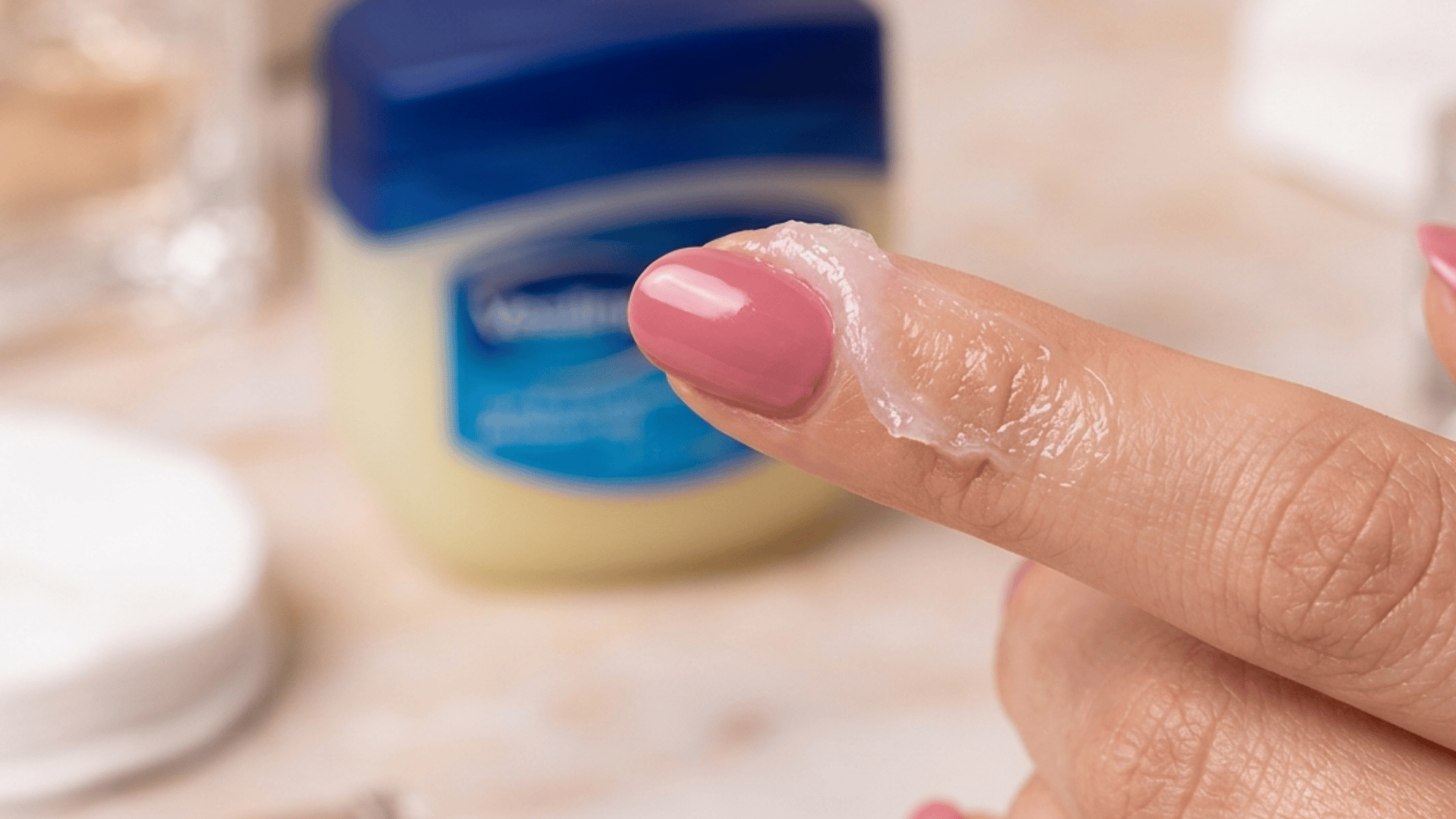Close-up of a fingertip with petroleum jelly applied, with a blurred Vaseline jar visible in the background