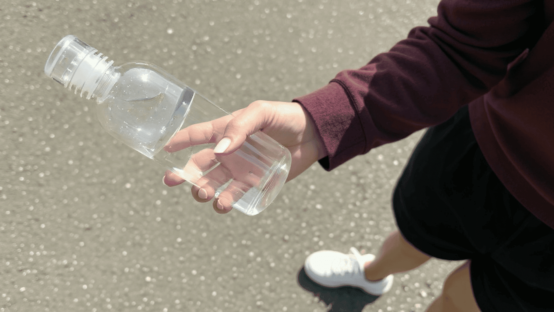 Person holding a clear plastic water bottle outdoors while walking on pavement in daylight