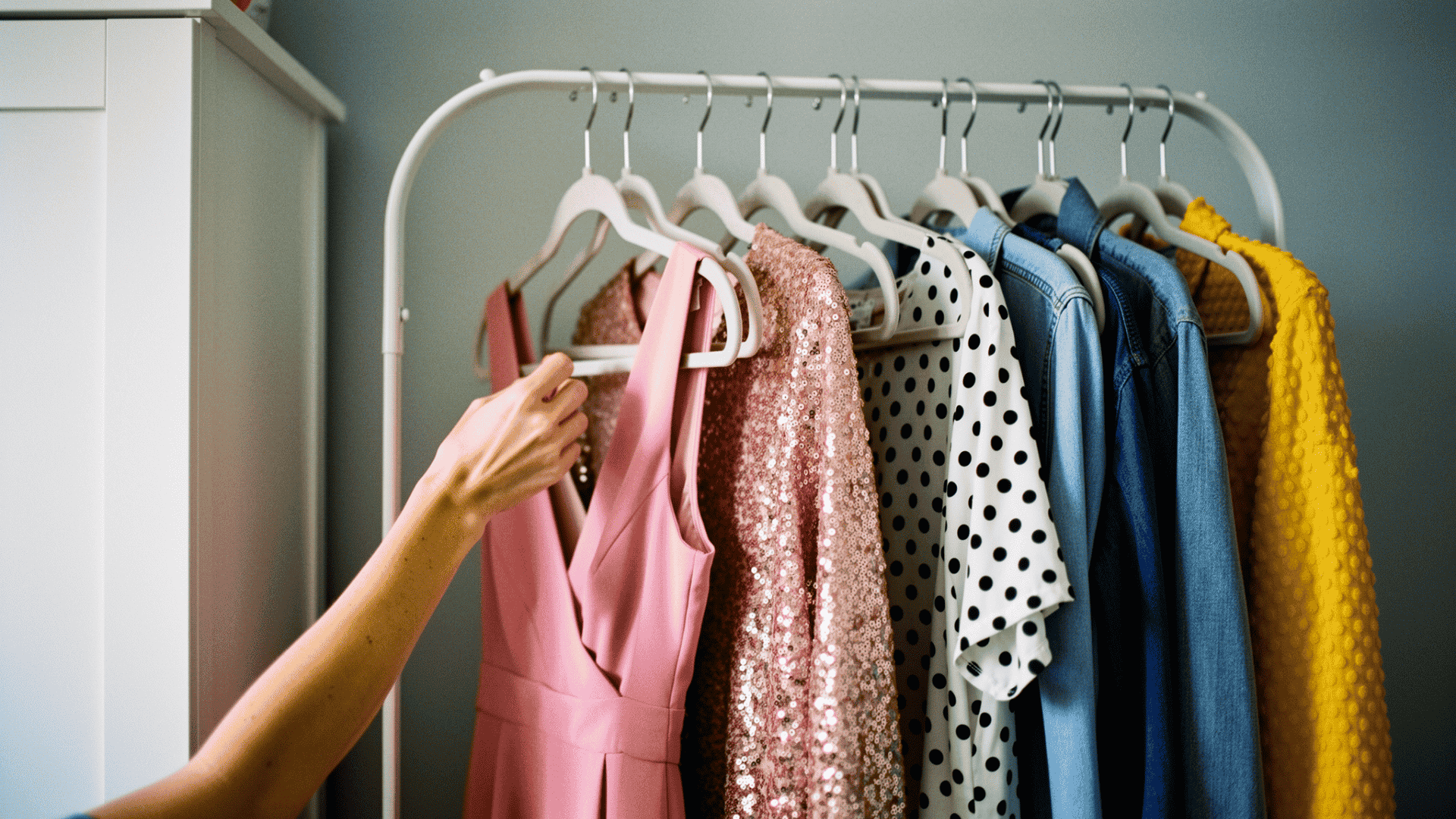 Person selecting clothes from a clothing rack with dresses, polka dot shirt, denim jacket, and yellow sweater