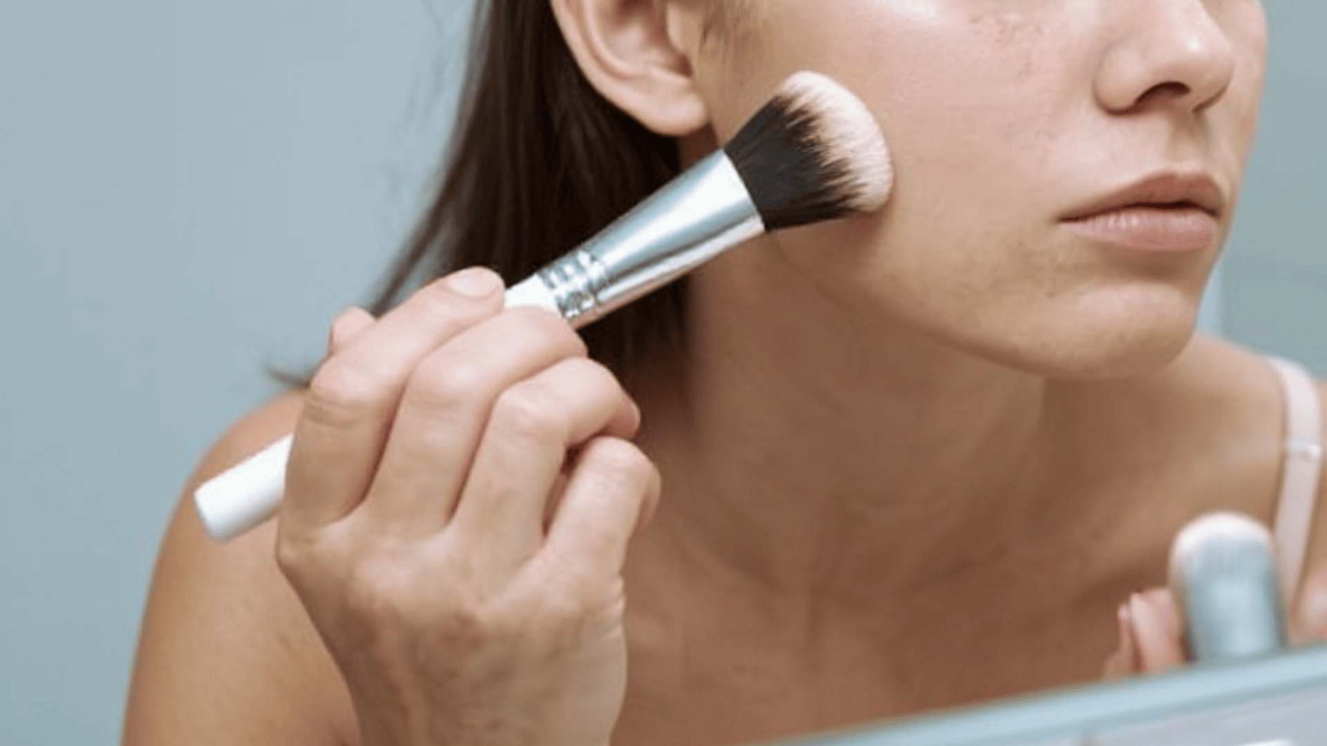 Woman applying foundation to her cheek using a makeup brush while looking in a mirror