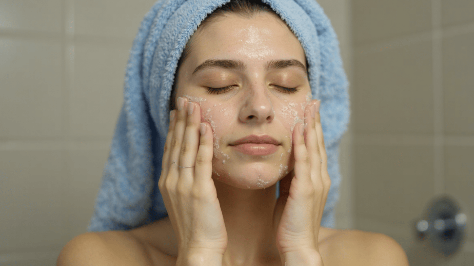 Woman gently exfoliating her face with a facial scrub while wearing a towel wrapped around her hair in a bathroom setting