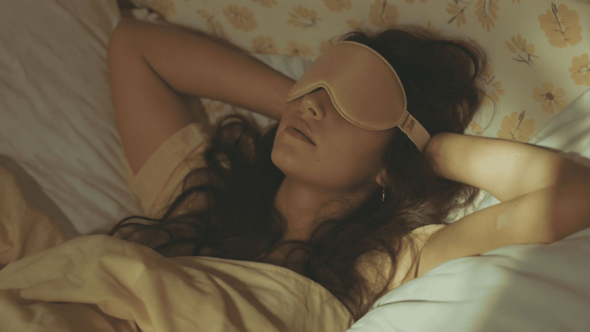 Woman lying in bed wearing a sleep mask, relaxing with hands behind her head on a pillow under soft warm bedroom lighting