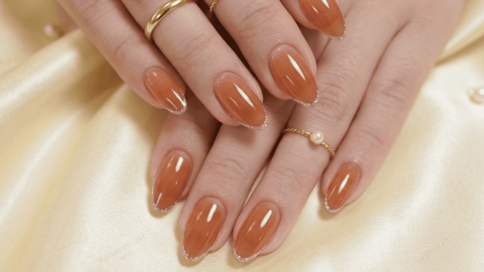 Close-up of almond-shaped nails with glossy brown polish and subtle glitter tips, styled with delicate gold rings