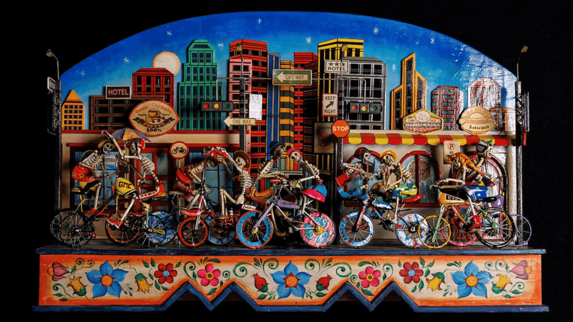 Colorful folk art diorama showing skeleton figures riding bicycles through a lively city scene with detailed buildings and decorative floral patterns