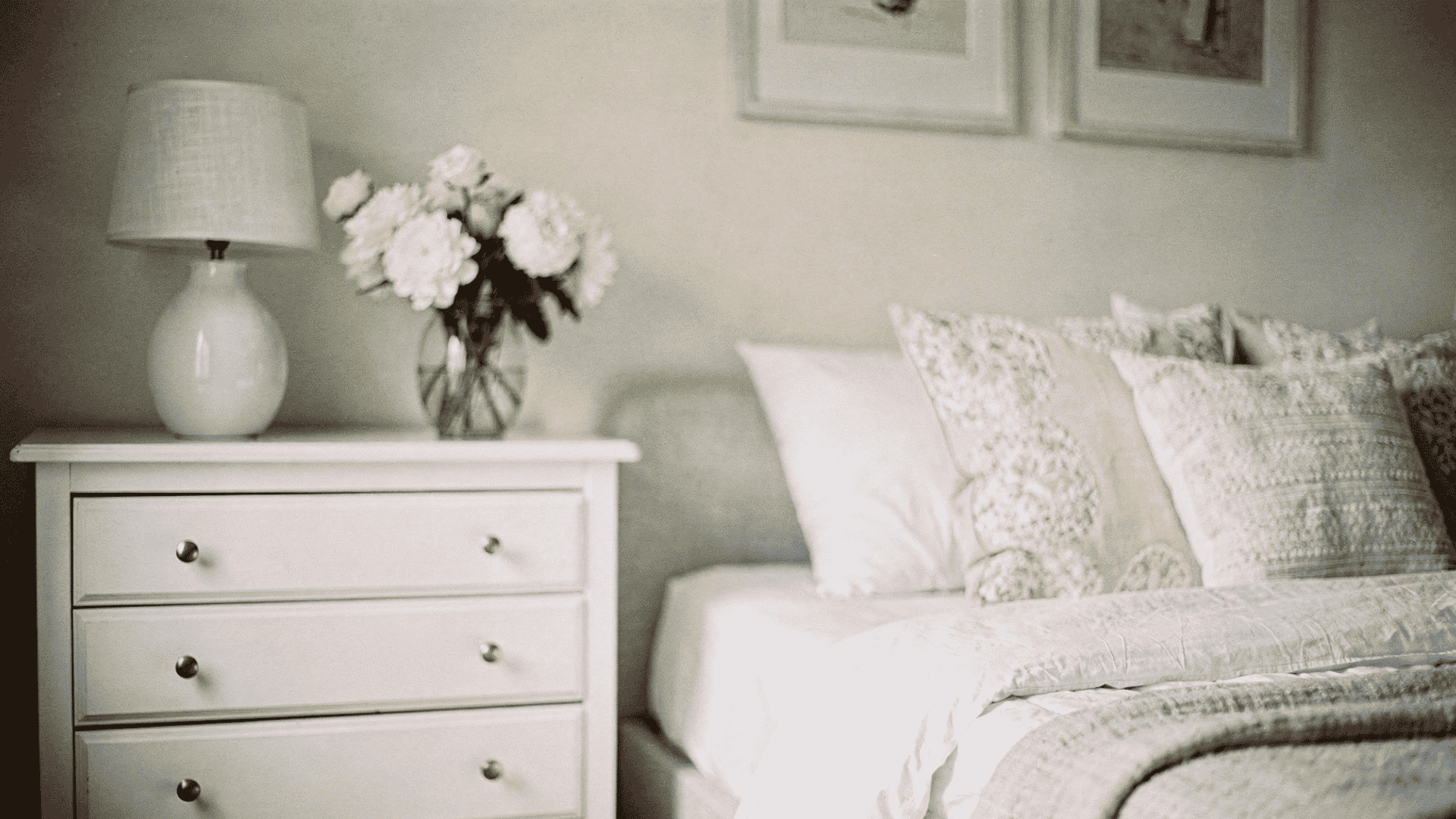 _Cozy bedroom corner with a white dresser, lamp, fresh flowers, and a bed with textured pillows in soft, neutral tones.