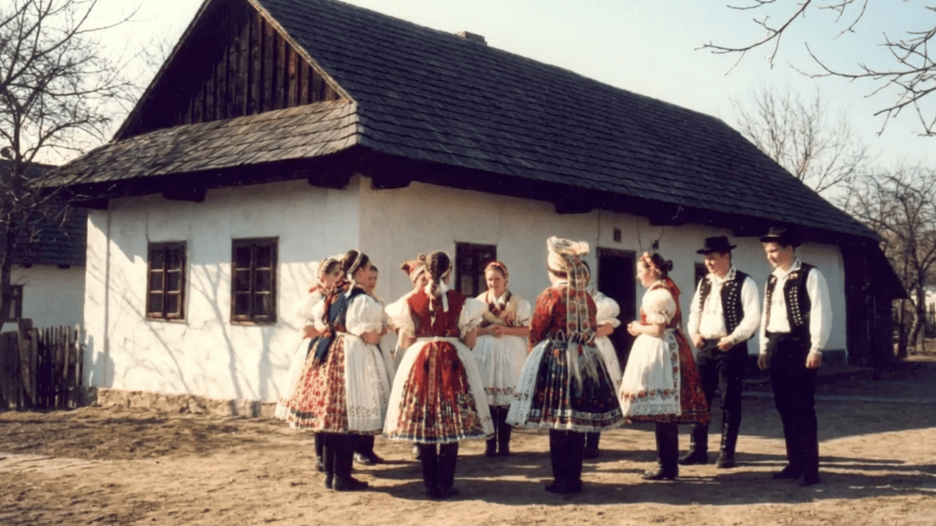 People in traditional folk costumes standing in a circle outside a rural house, reflecting cultural customs and community traditions