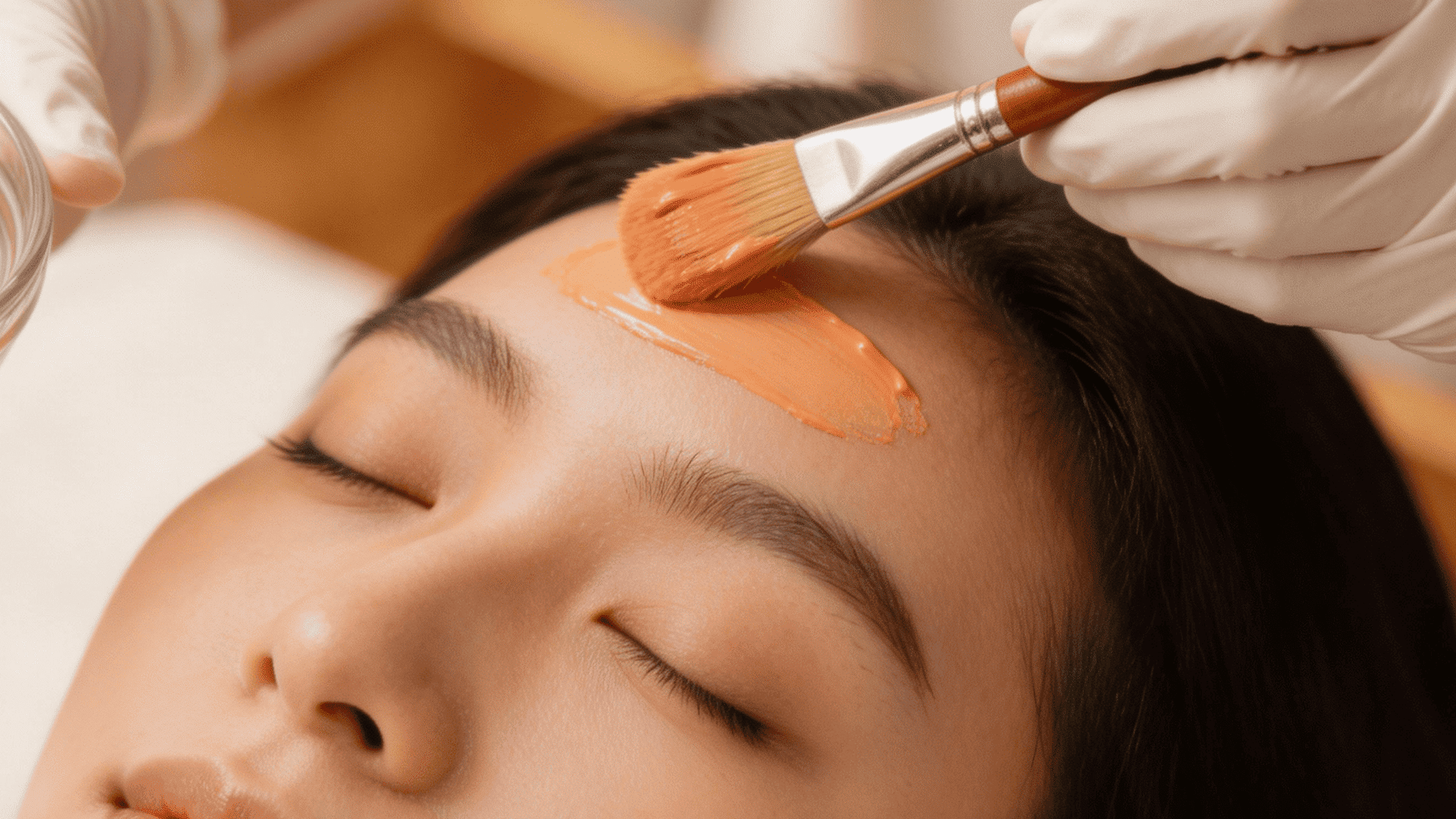 Person applying an orange facial mask to a womanโs forehead using a brush during a spa treatmen