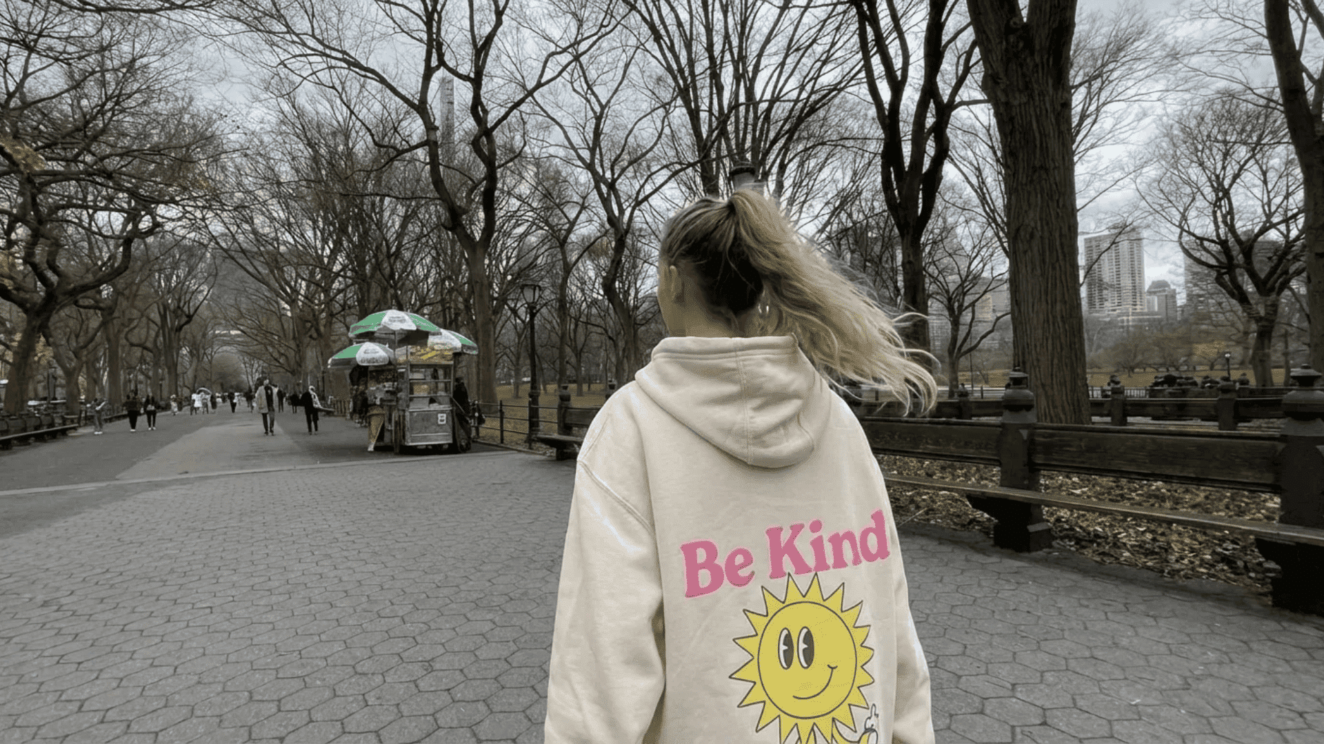 Person in a beige hoodie with “Be Kind” and a smiley graphic walking through a park with bare trees and city buildings in the background