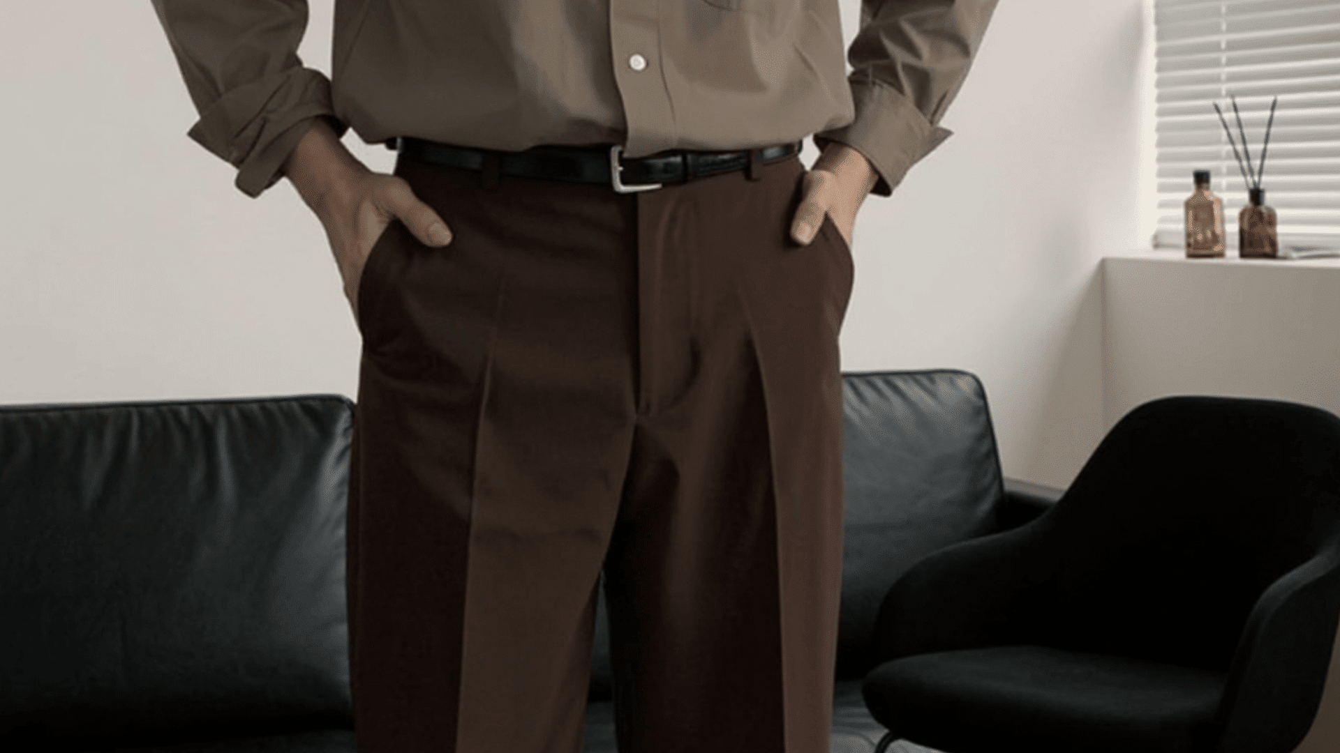 Person wearing brown trousers and a tucked-in shirt standing indoors with hands in pockets near a sofa