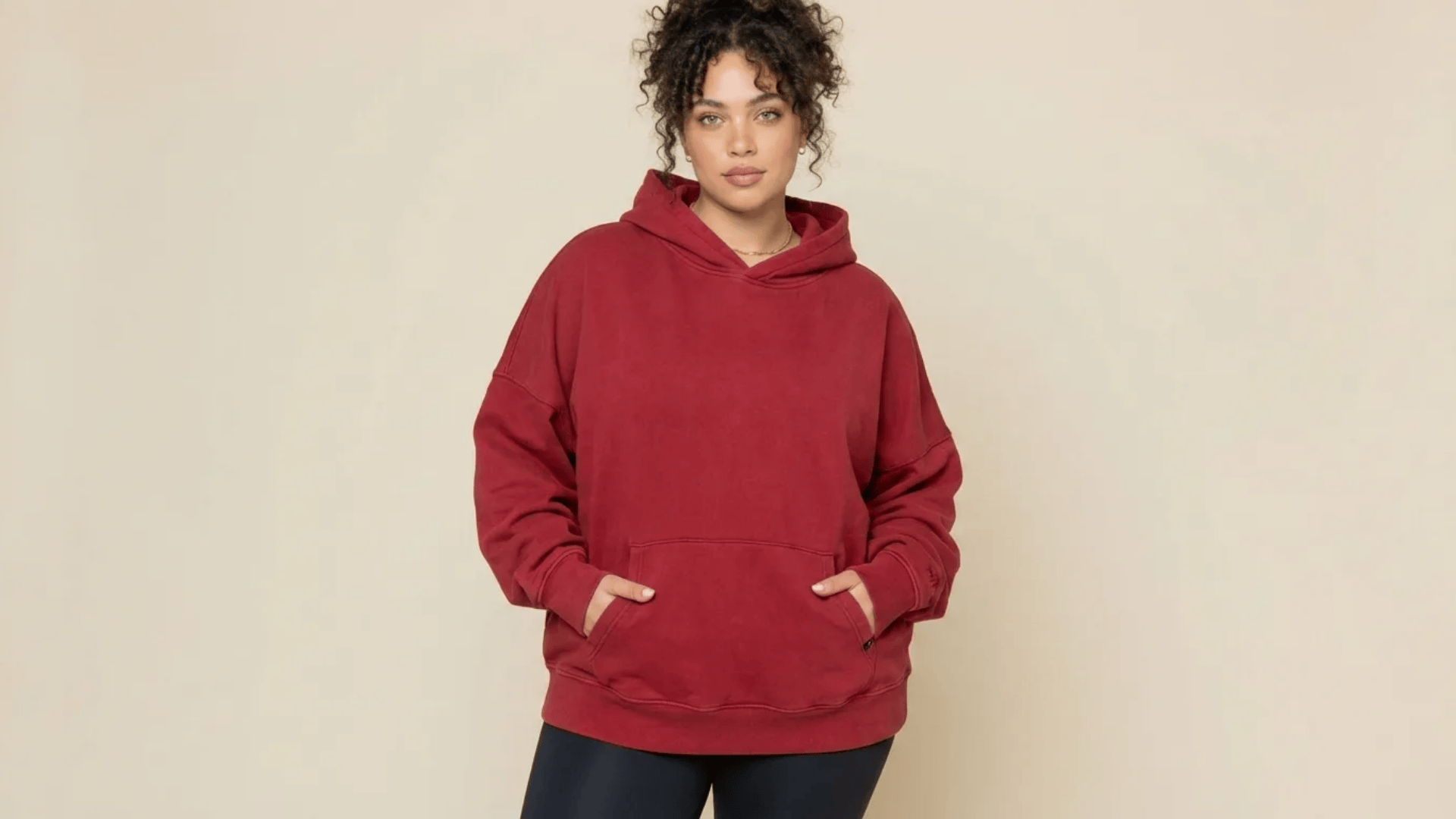 Woman wearing a red hoodie with front pocket, standing against a plain beige background.