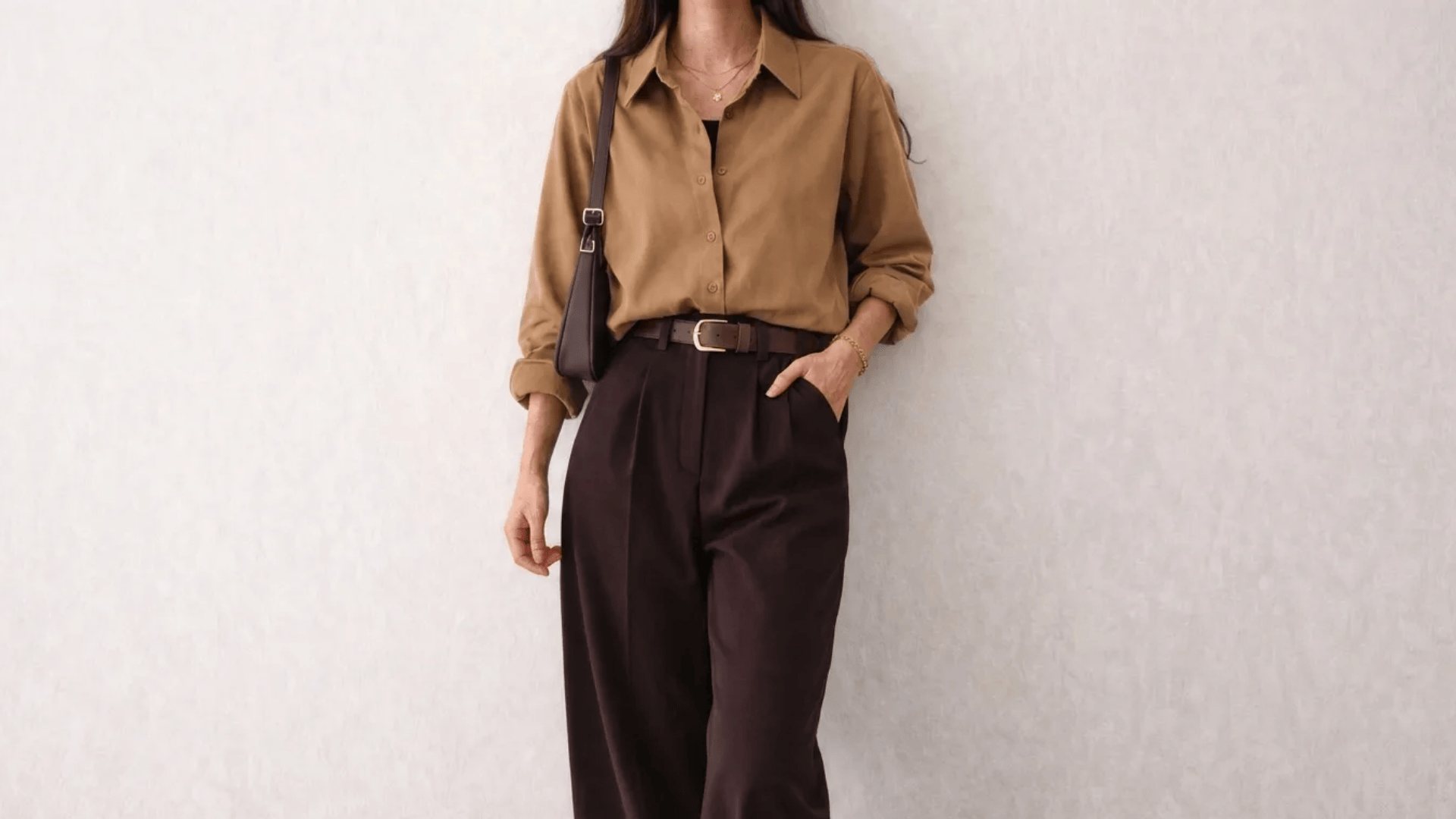 Woman wearing a tan button-up shirt with dark brown trousers, a matching belt, and a brown shoulder bag against a neutral wall