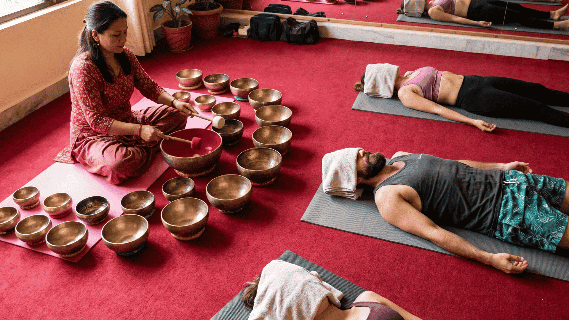A woman plays Tibetan singing bowls for relaxed participants during a sound healing session in a bright studio