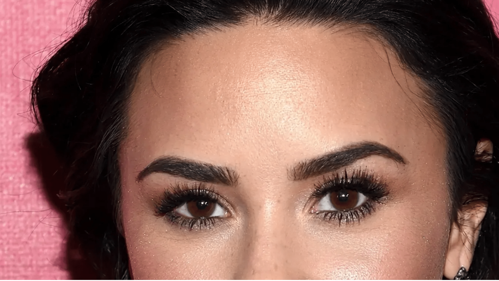 Close-up of brown eyes with bold lashes, defined eyebrows, glowing skin, and dark hair against a pink background.
