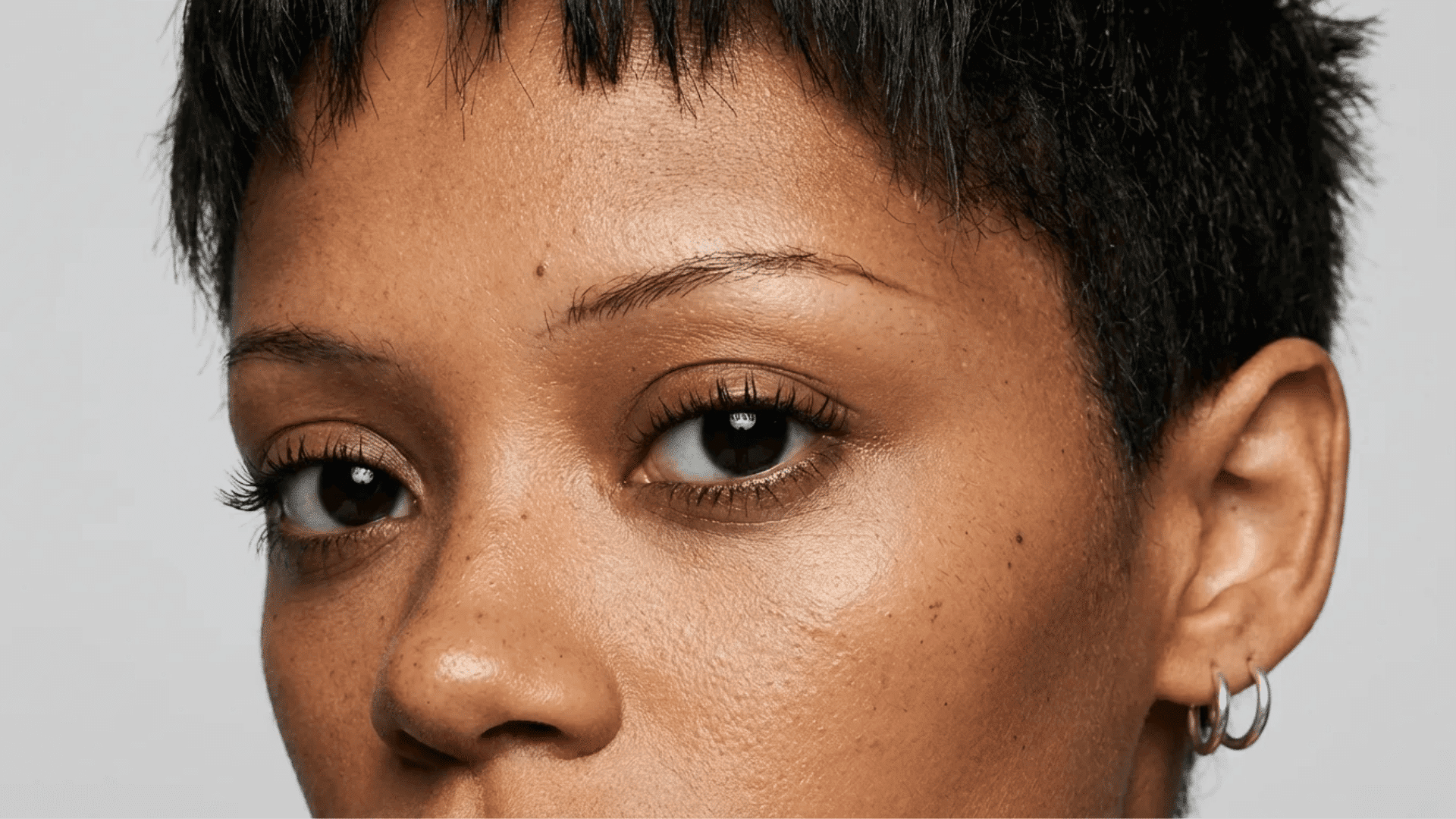 Close-up portrait of a person with short dark hair, brown eyes, natural skin texture, and small hoop earrings, looking slightly to the side.