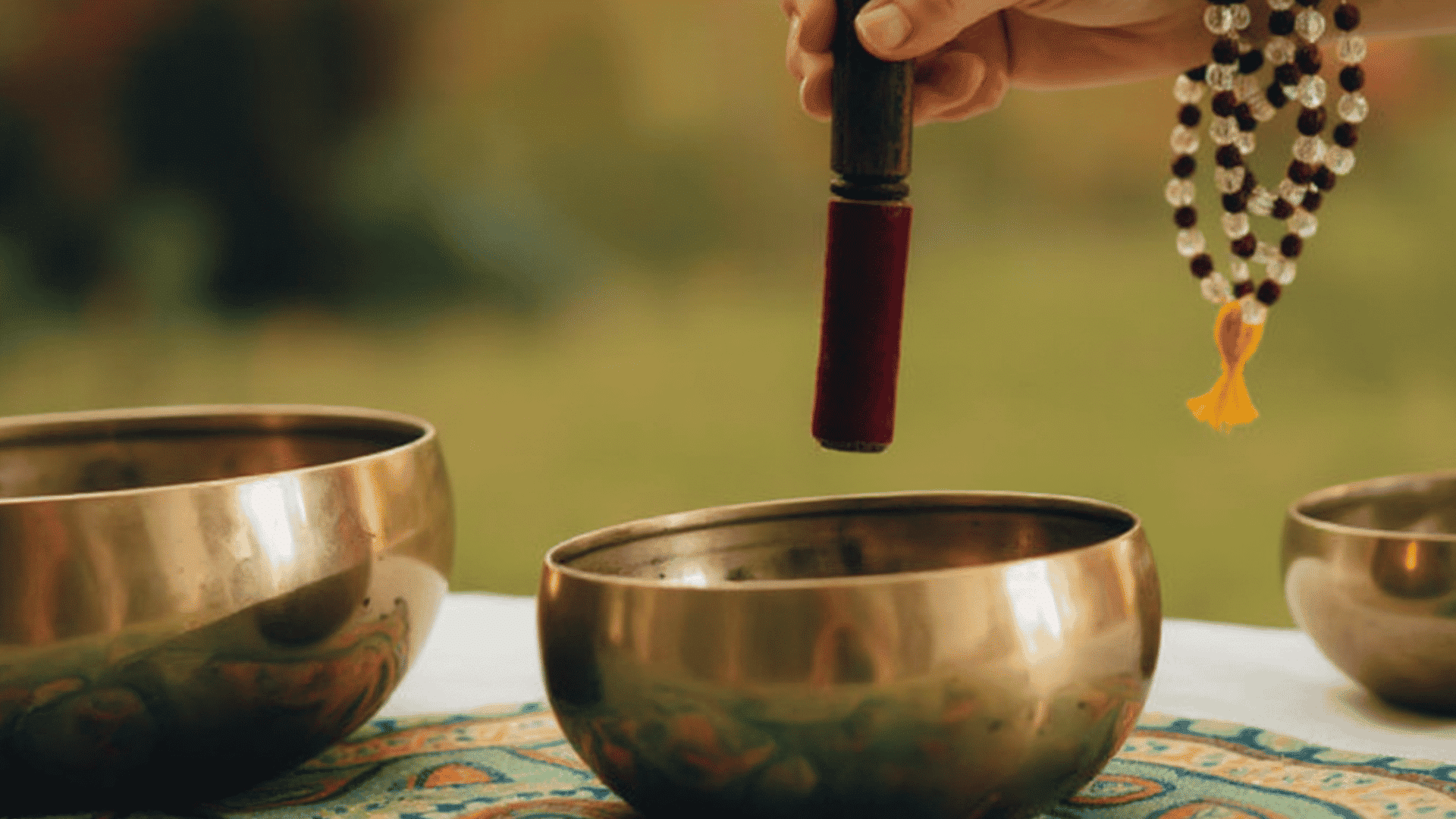 Hand holding a mallet over metal bowls. Explaining what is sound healing with tools in a serene setting.