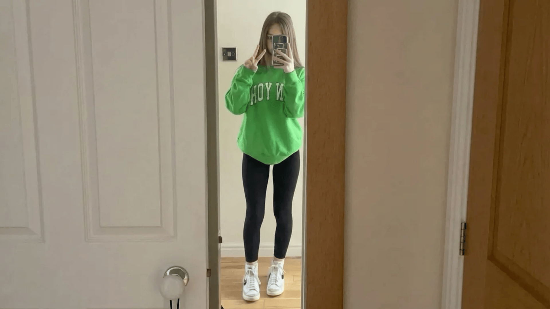 Person in mirror selfie wearing oversized green sweatshirt, black leggings, and white sneakers, holding phone and making peace sign (1)