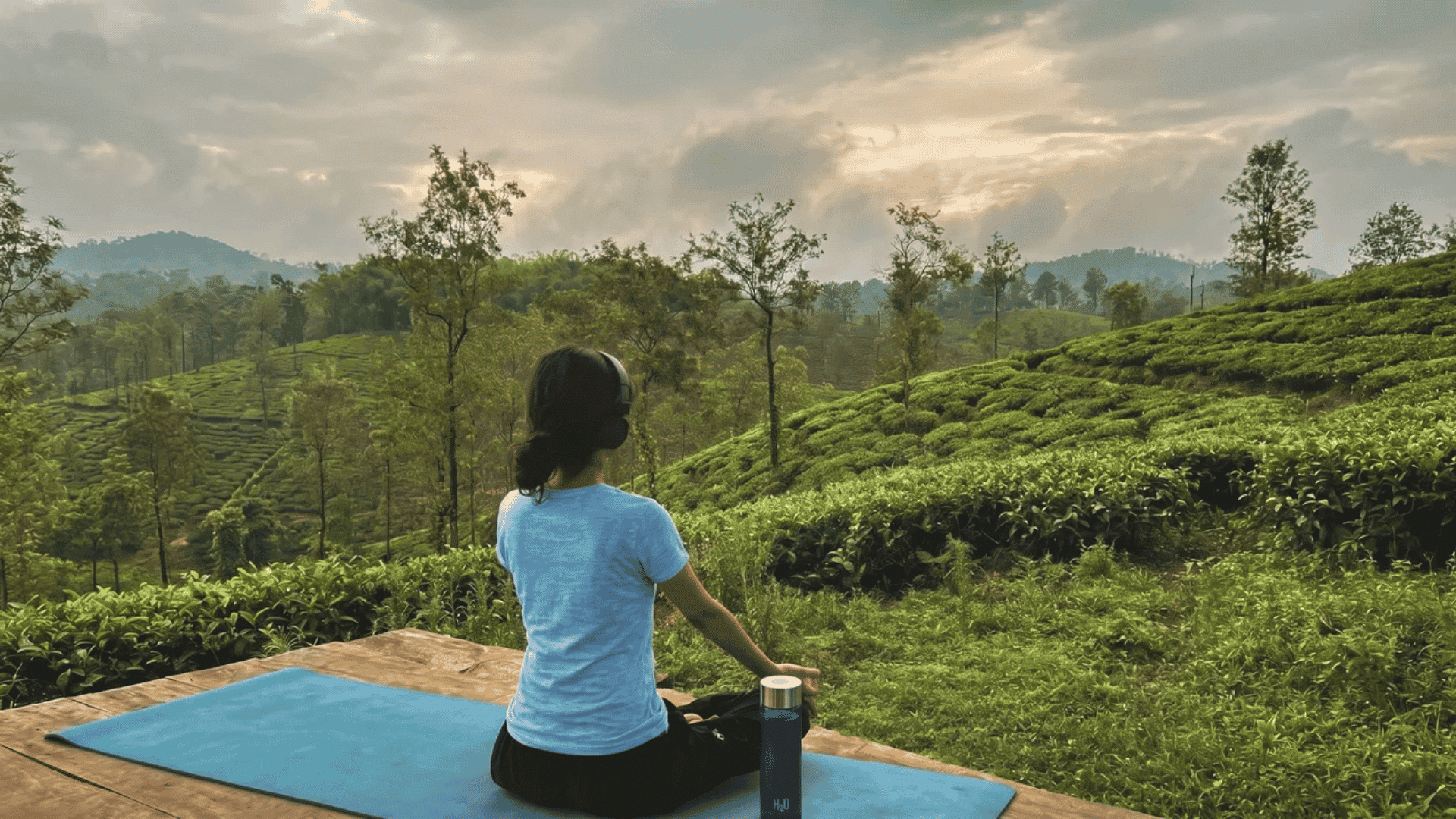 Person sitting cross-legged on a yoga mat, wearing headphones, overlooking lush green hills in a calm outdoor meditation setting.