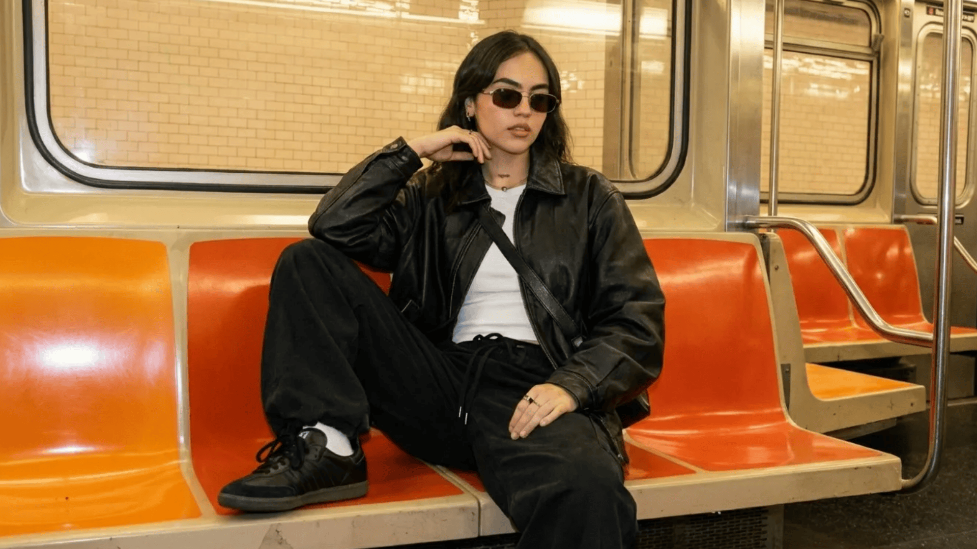 Person wearing a black leather jacket, white tee, and black pants, sitting on an orange subway seat with sunglasses.