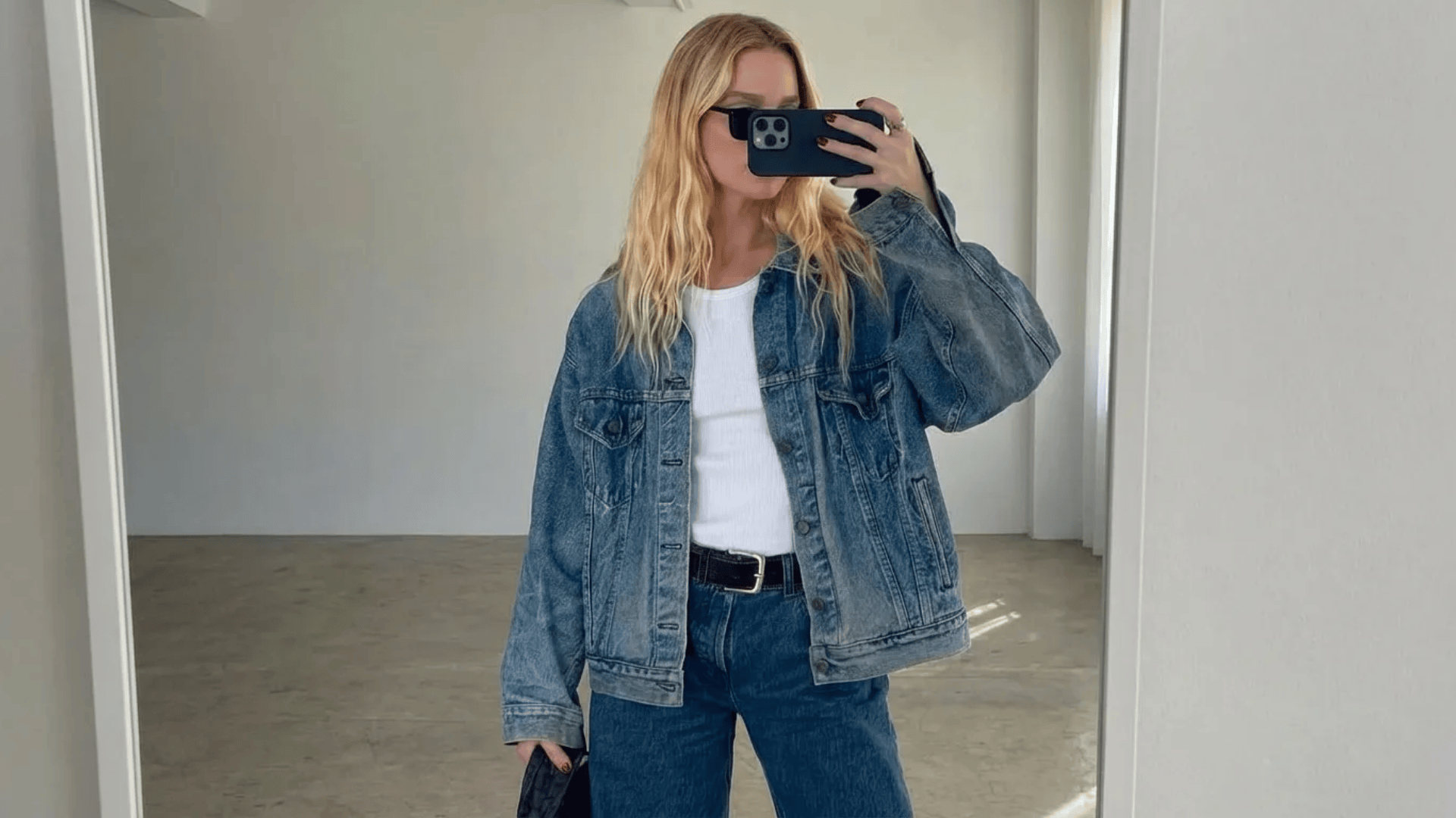 Person wearing a denim jacket over a graphic tee with blue jeans and a belt, taking a mirror selfie indoors. (1)