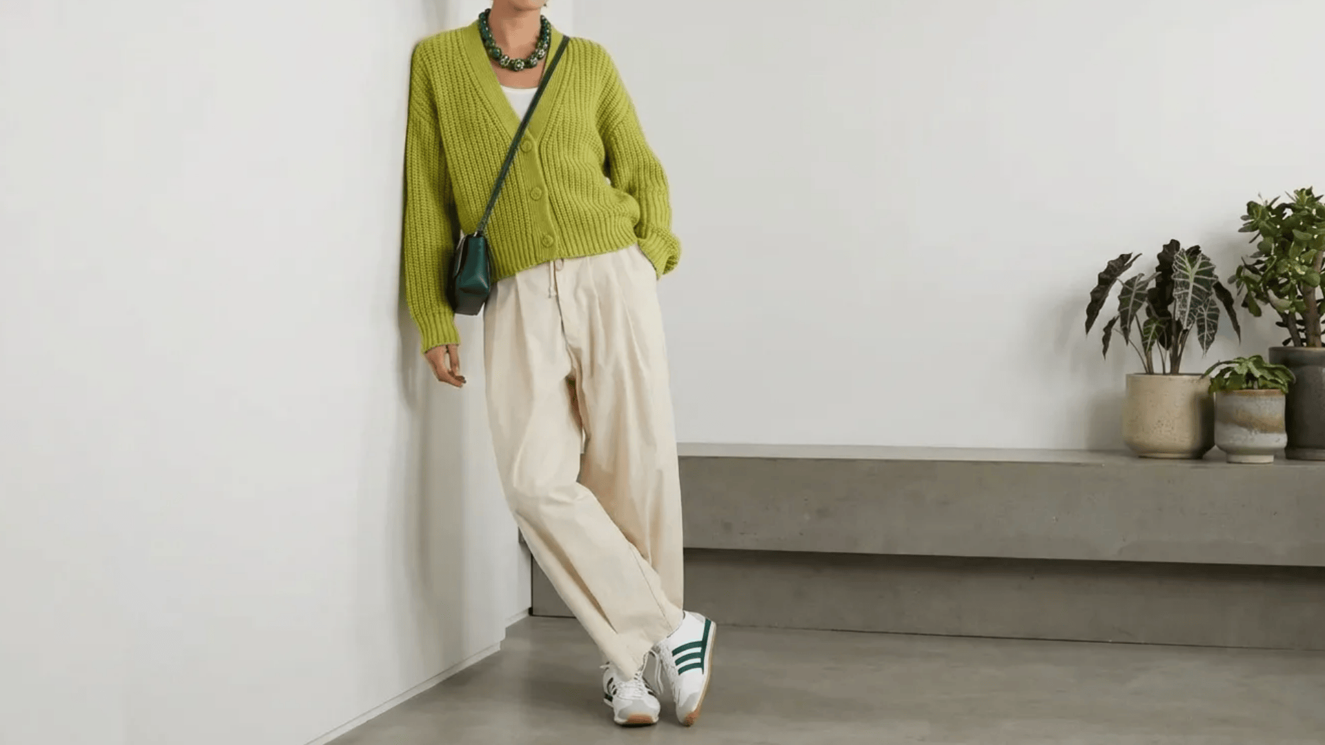 Person wearing a green cardigan with cream pants and sneakers, leaning against a wall with a crossbody bag and necklace.