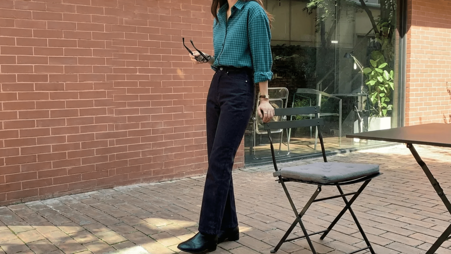 Person wearing a green checked shirt with high-waist dark jeans and black boots, standing outdoors near a brick wall and chair.