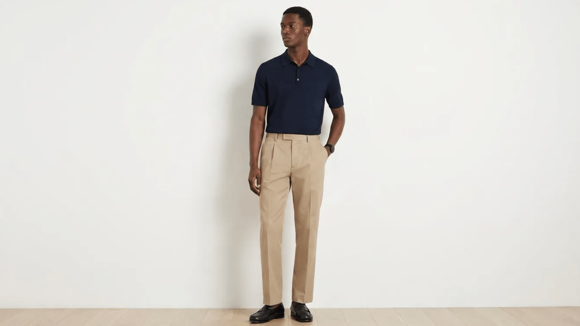 Person wearing a navy polo shirt with beige trousers and black shoes, standing against a plain wall.