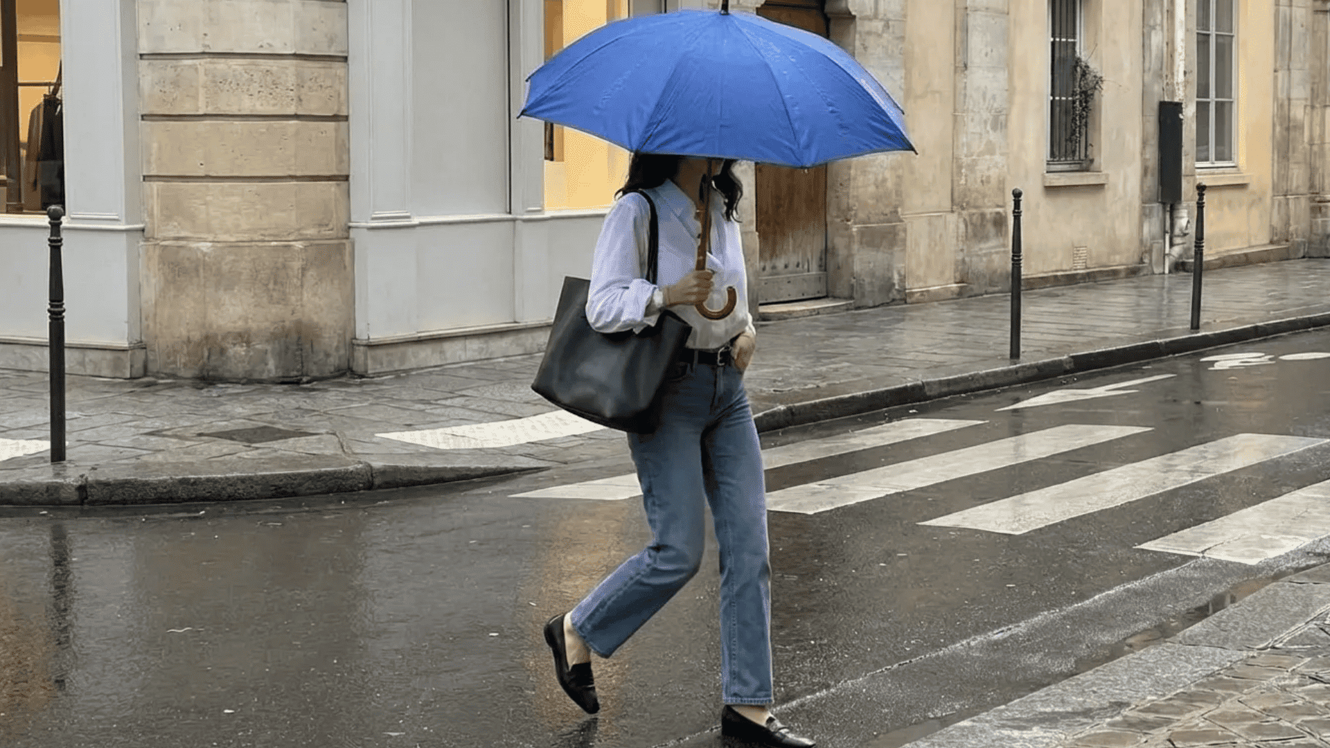 Person wearing a white shirt with blue jeans and black shoes, walking in the rain with a blue umbrella on a city street.