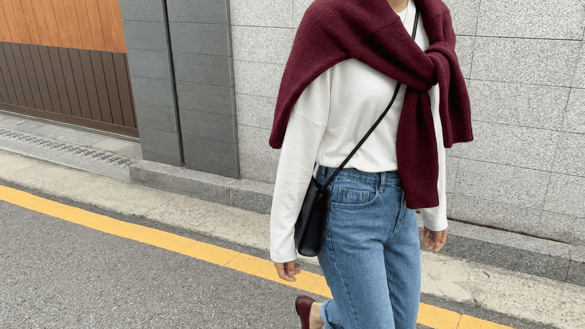 Person wearing a white top with blue jeans and a sweater tied around the shoulders, walking on a street with a crossbody bag.