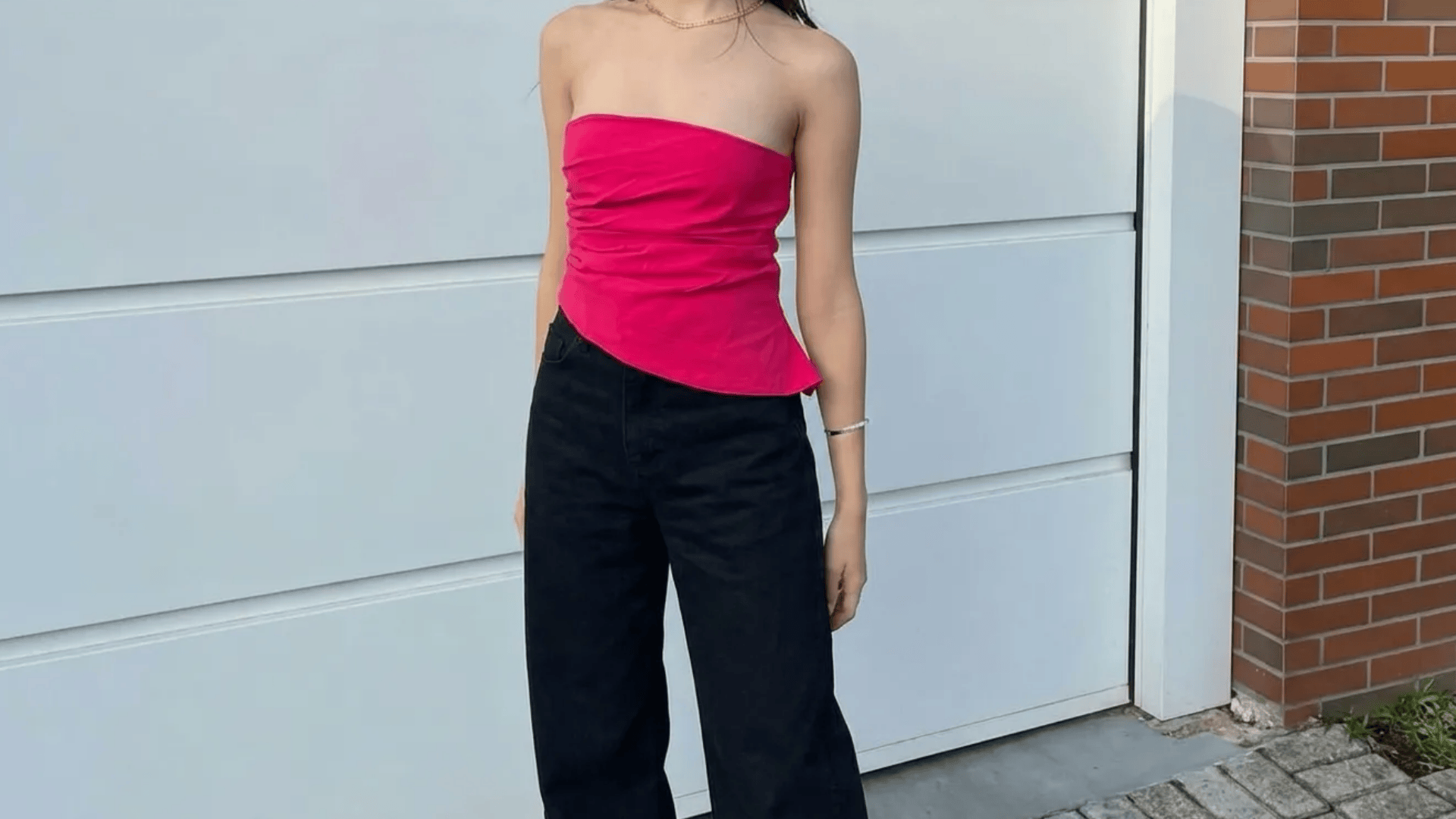 Person wearing bright pink strapless top with black wide-leg pants, standing outdoors near a wall