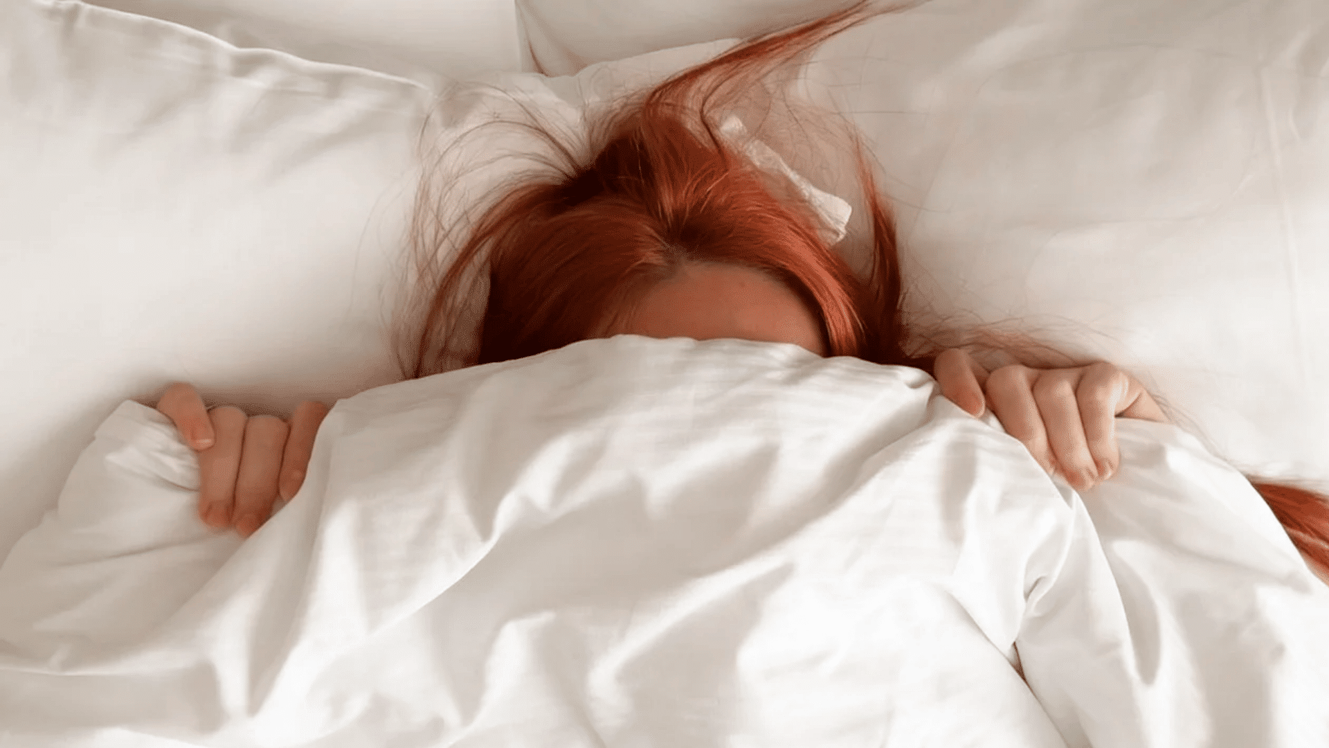 Person with red hair lying in bed, hiding under white blankets with only the top of their head and hands visible, creating a cozy, sleepy mood.