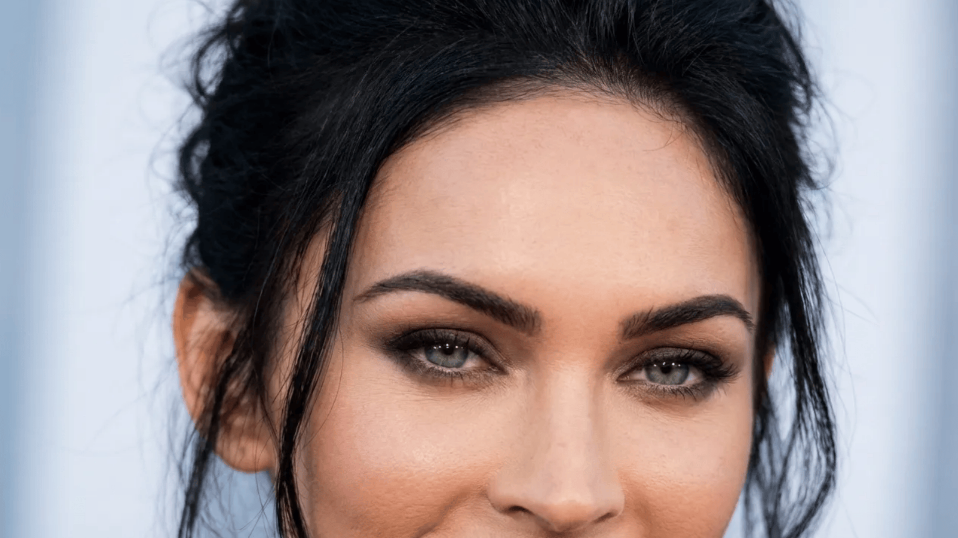 Portrait of a woman with green eyes, dark hair styled up, defined eyebrows, and soft natural makeup, looking directly at the camera.