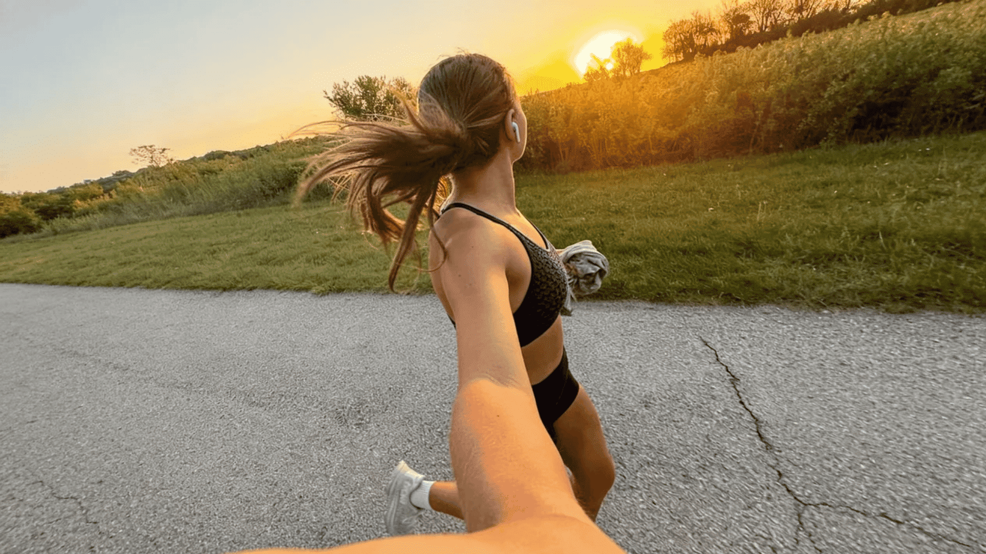 Woman jogging outdoors at sunset, captured in a dynamic selfie angle with hair flowing and golden light over a scenic path.