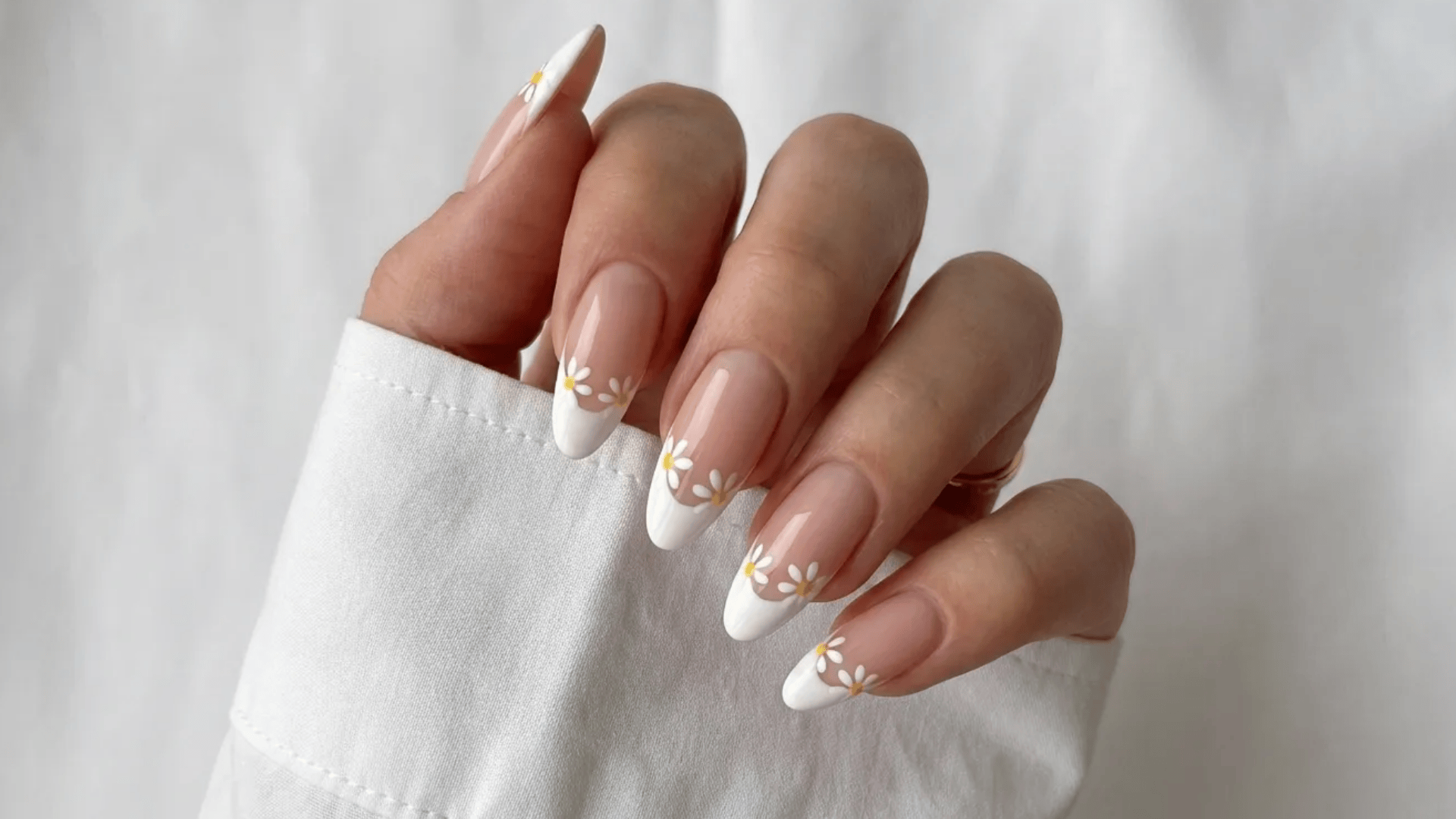 Almond-shaped nude nails with white French tips and delicate daisy flower nail art, hand partially covered with a white sleeve.