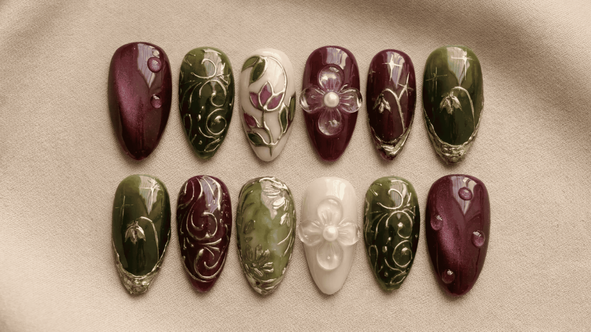 Assorted press-on nail designs in burgundy, green, and nude tones with gold detailing, floral patterns, and 3D embellishments displayed on a neutral fabric background.