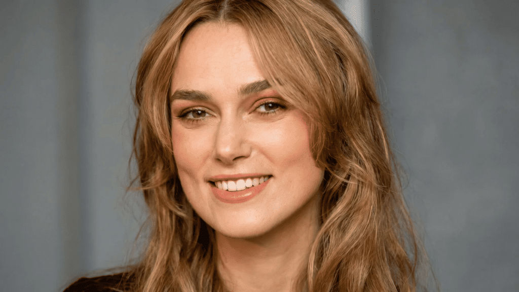 Close-up of a smiling woman with warm-toned honey blonde hair in soft waves, natural glowing makeup, and a neutral blurred background.