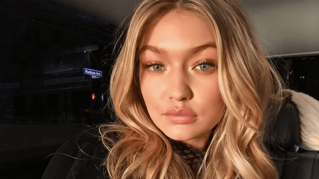 Close-up of a woman with warm-toned golden blonde hair and soft waves, glowing skin, and natural makeup, sitting in a dimly lit car with a blurred city background.