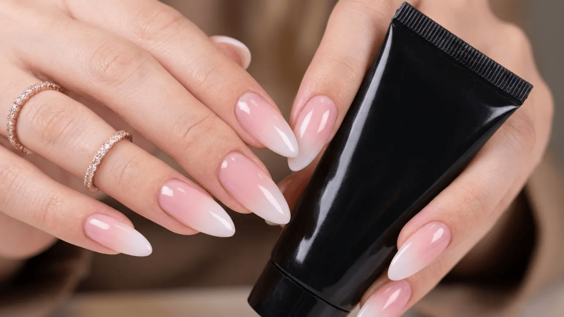 Glossy ombré pink to white almond-shaped nails, hands holding a black cosmetic tube with delicate gold rings.