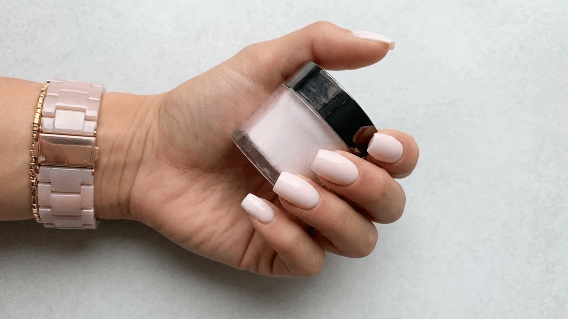 Glossy pale pink manicure on medium square-shaped nails, hand holding a matching nail polish bottle with a soft neutral background.