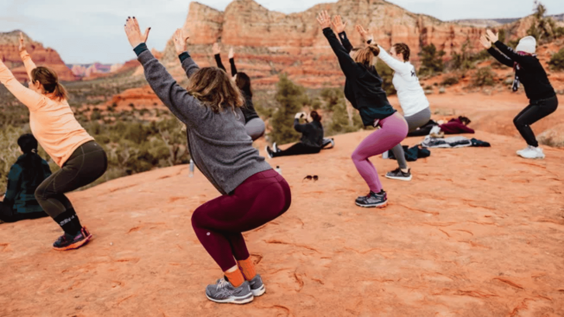 Group of people doing a squat exercise outdoors on a rocky desert landscape, with red rock formations and hills in the background.
