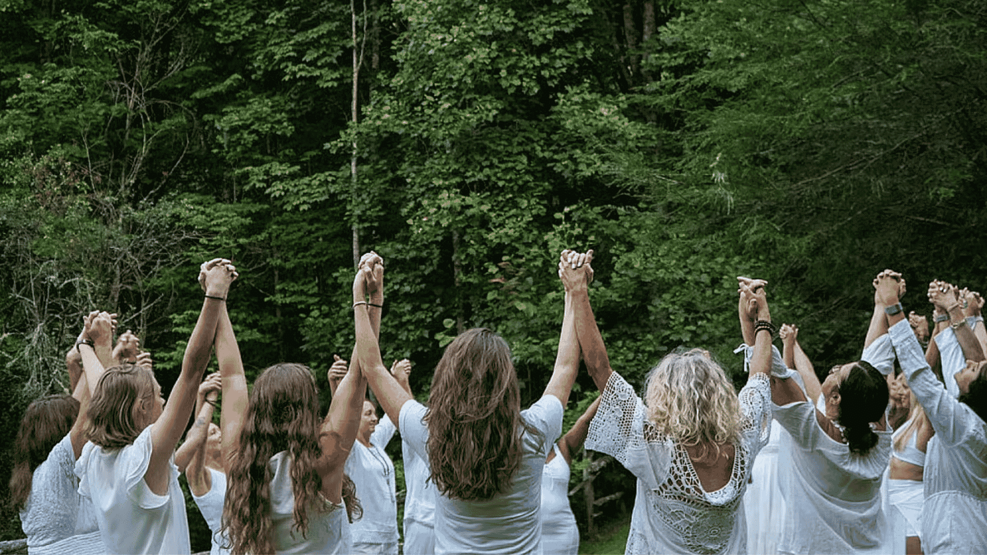 Group of women standing in a circle outdoors, holding hands up together in a forest setting, creating a sense of connection and shared energy.