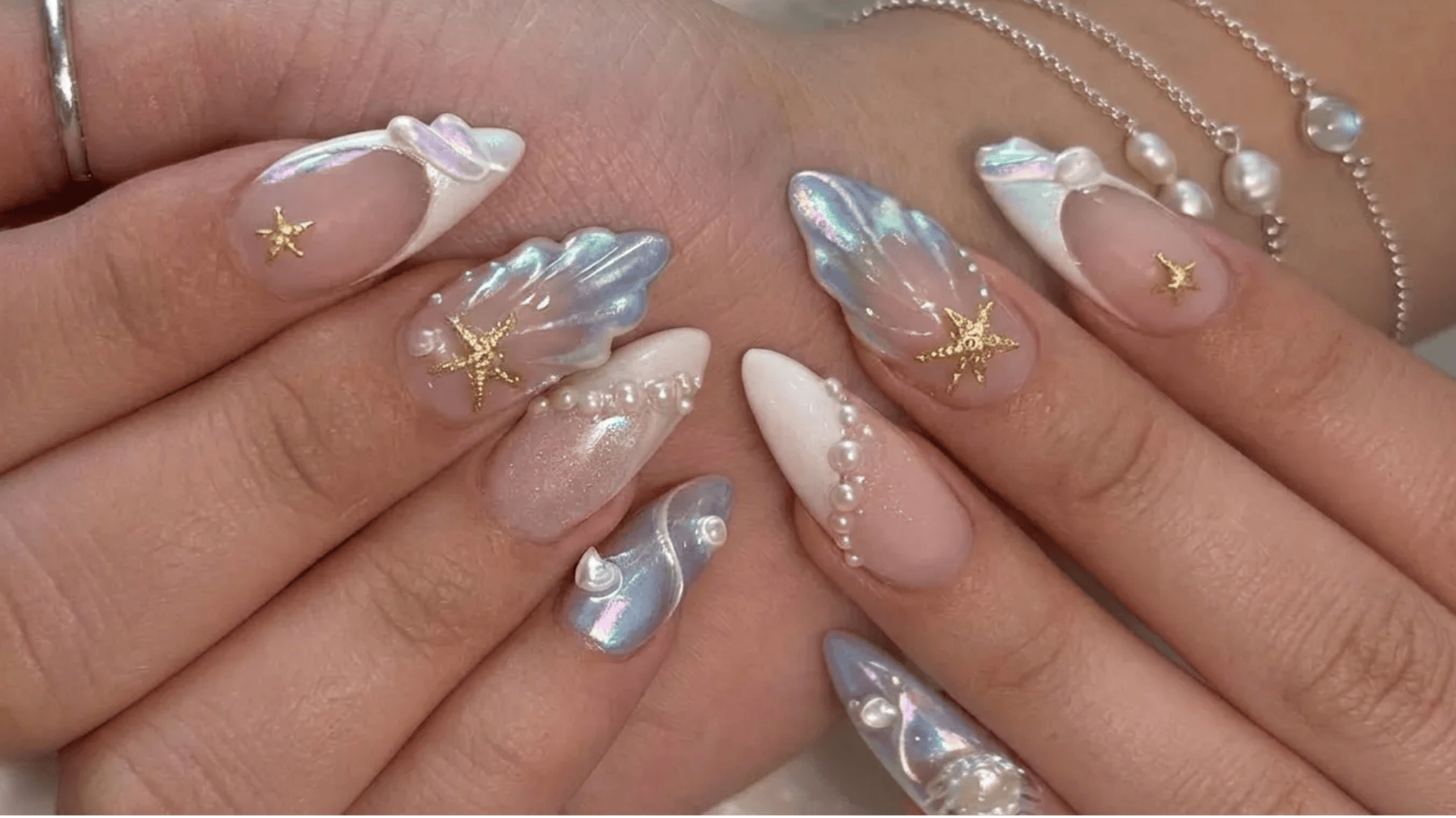 Long almond and stiletto nails with iridescent chrome finish, 3D shell designs, pearls, and gold star embellishments on a soft nude base.