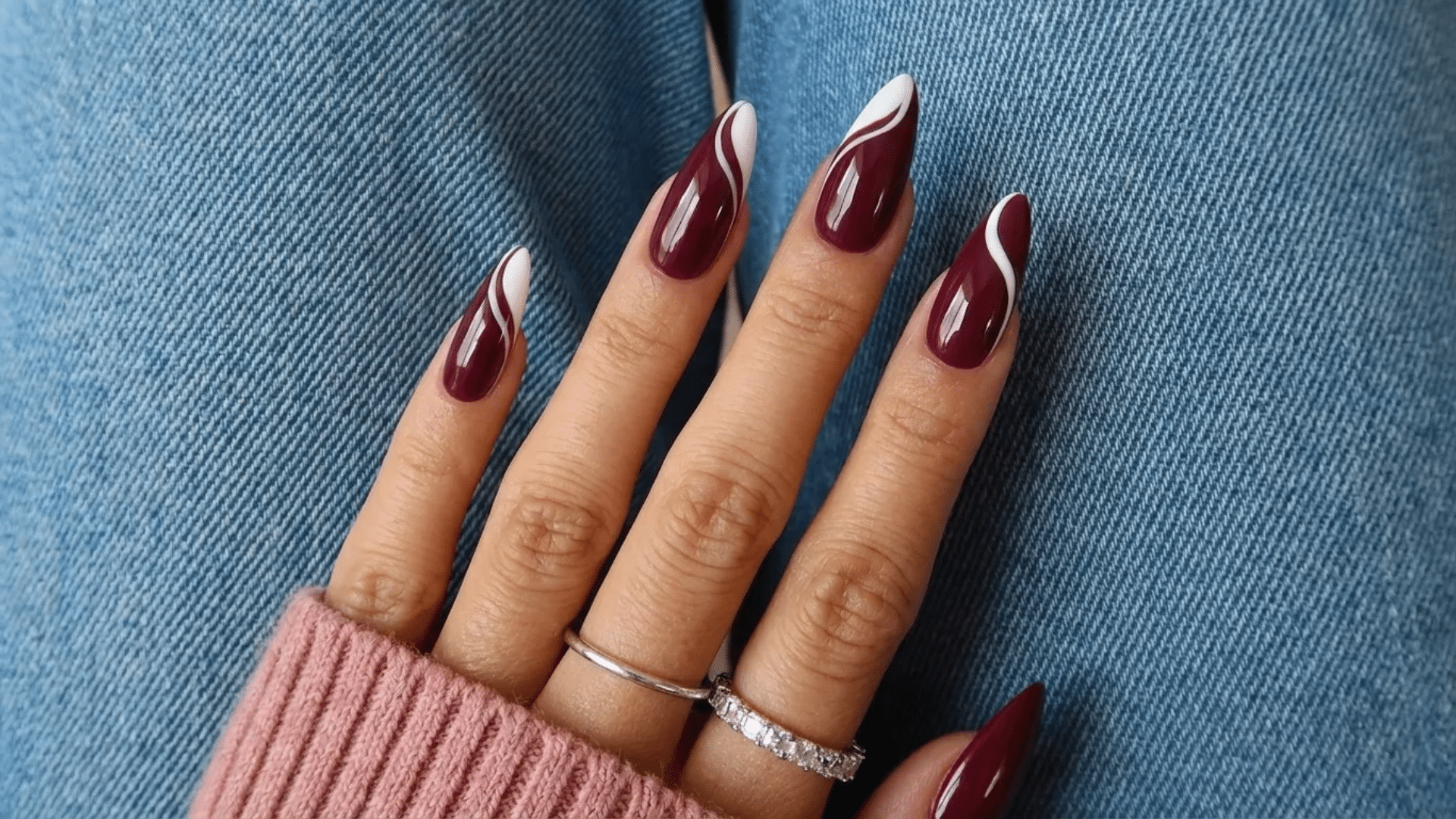Long almond-shaped nails with deep burgundy polish and white swirl accents, styled on a hand resting against denim fabric.