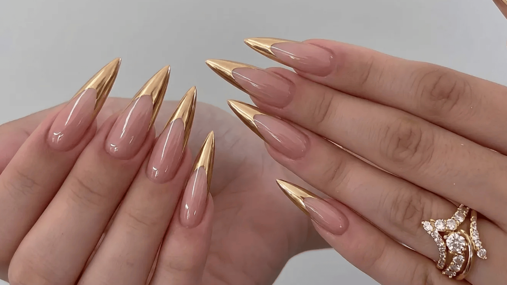 Long stiletto-shaped nude nails with metallic gold tips, styled on both hands with glossy finish and statement rings.