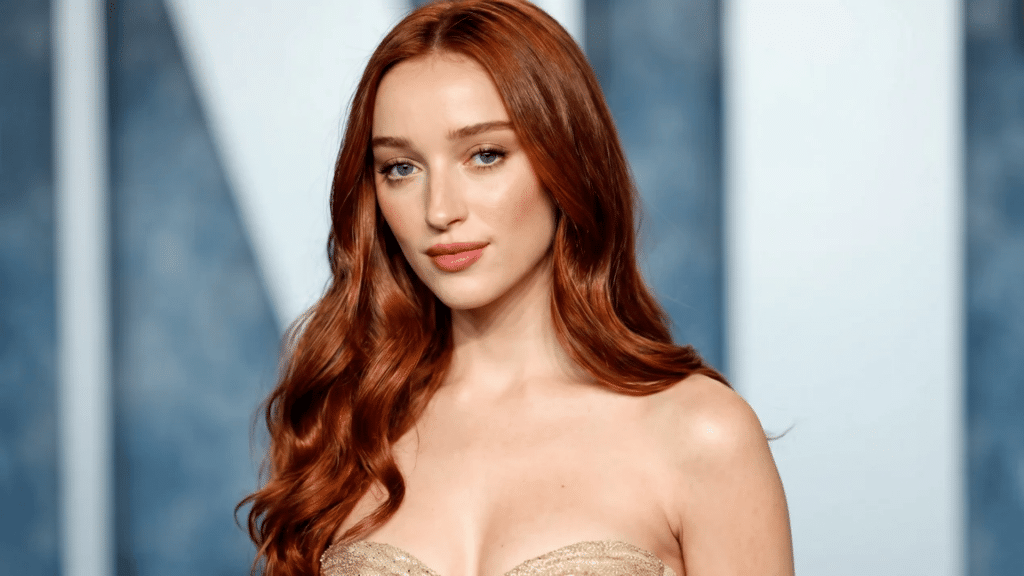 Portrait of a woman with long, wavy copper-red hair, warm glowing skin, and soft makeup, wearing a strapless gold outfit against a blurred neutral background.