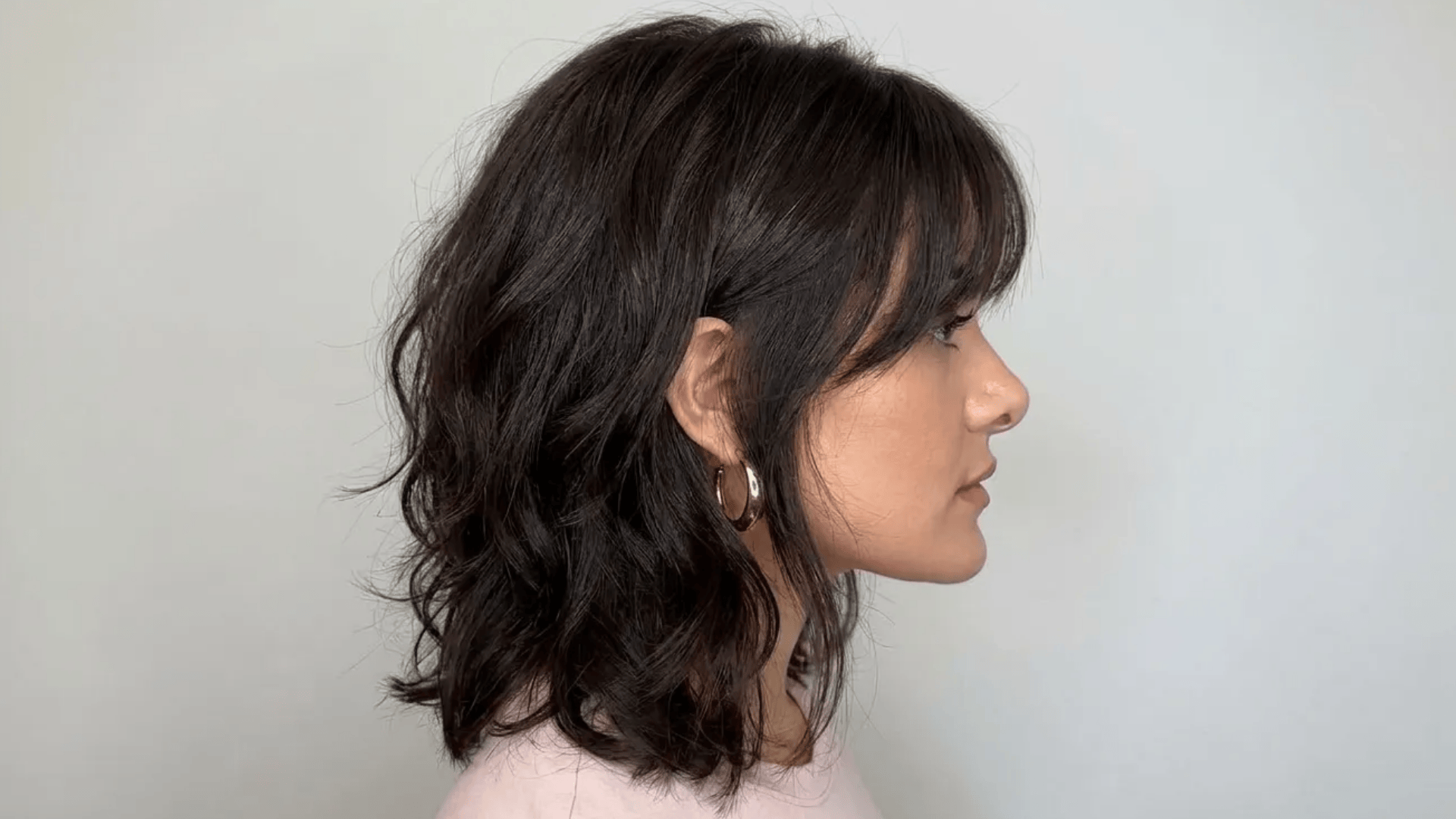 Side profile of a woman with shoulder-length layered wavy dark hair and bangs, wearing a gold hoop earring against a plain background.