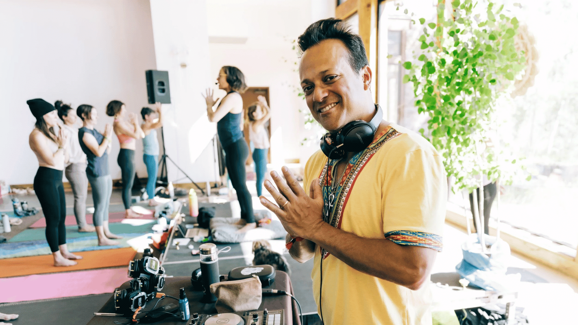 Smiling instructor with headphones leading a yoga class indoors, while a group of women practice poses on mats in a bright studio.