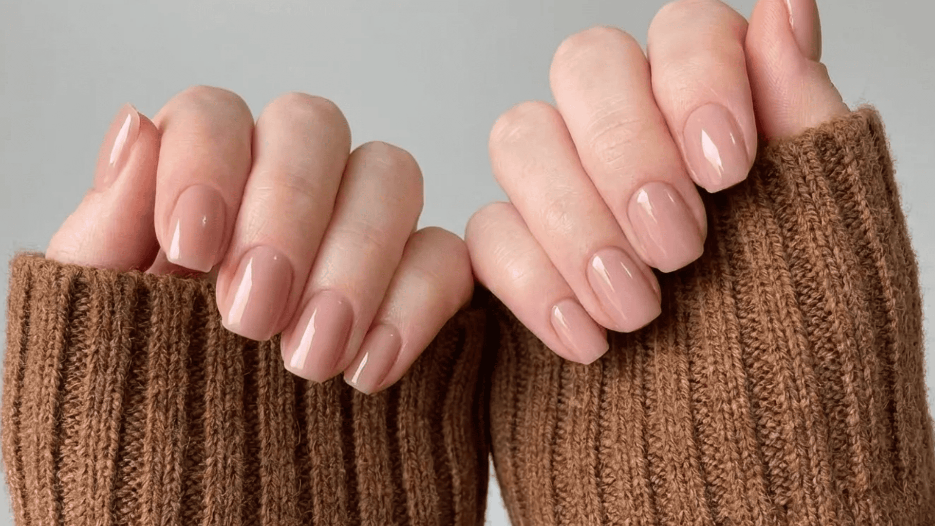 Soft nude gel manicure on short, rounded nails with glossy finish, hands partially covered in a brown knitted sweater.