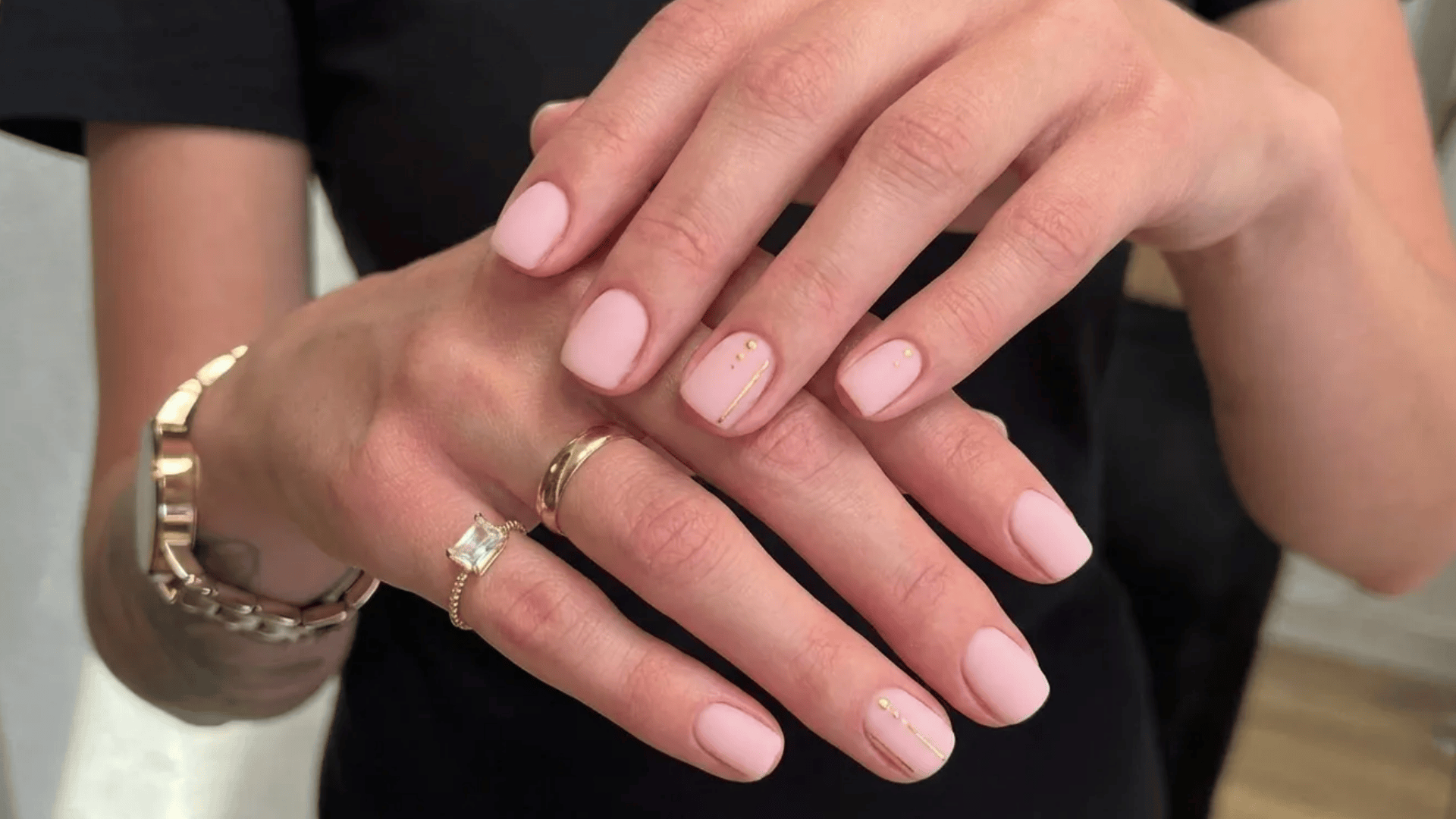 Soft pink matte manicure on short oval nails with minimal gold line and dot accents, hands styled with gold rings and bracelet.