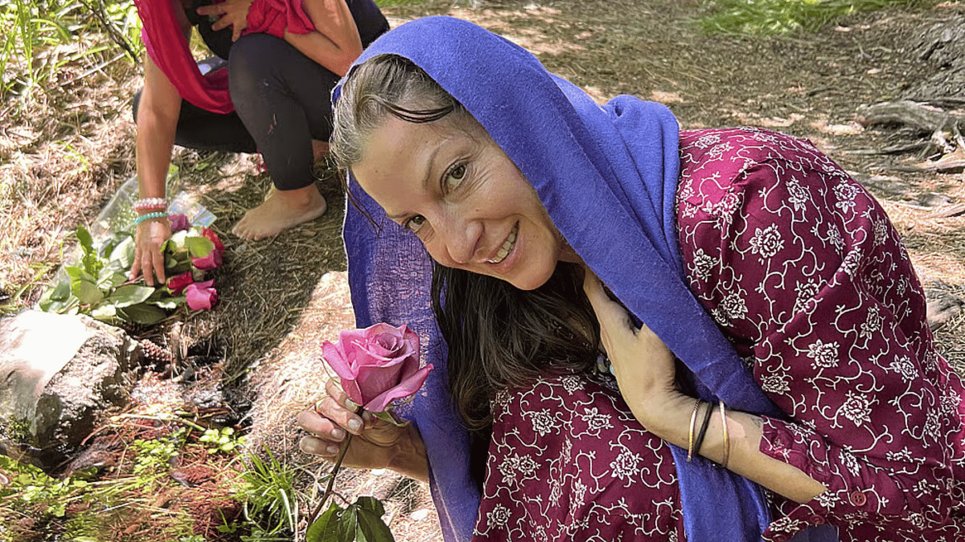 Woman in a nature setting holding a pink rose near a small stream, with others in the background placing flowers, creating a calm and reflective retreat moment.