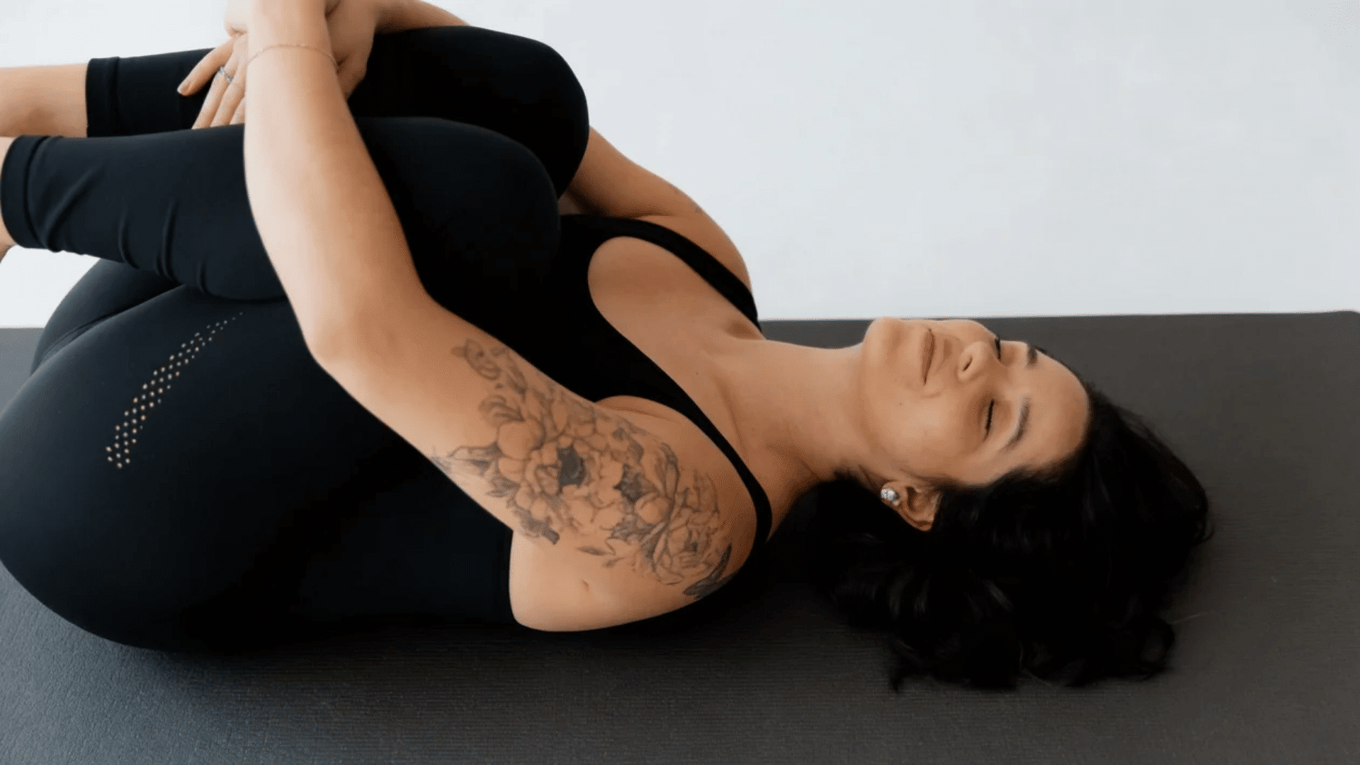 Woman lying on her back performing Knees-to-Chest Pose (Apanasana), gently hugging both knees toward her chest with eyes closed on a yoga mat in a calm, minimal setting.